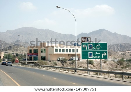 On a freeway, a sign indicates Dubai and Oman. A pharmacy, with trucks on its parking, stands close to the road. There are some cars on the road and arid mountains are visible in the background.
