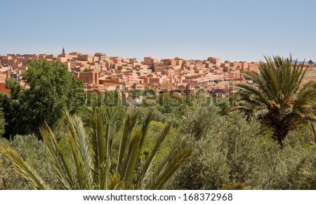 The town of Boumalne appears on the top of a hill under a blue sky. Palm trees and olive trees grow in the foreground in the valley.