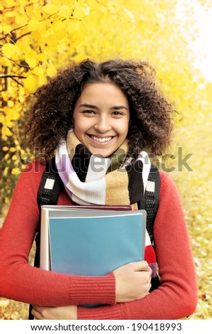 A smiling cute young female student with books in hand on the background of the autumn landscape. Female student in warm autumn.