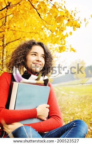 Female student in warm autumn. A cute Latino American young girl with books in hand sitting on the background of the autumn landscape.