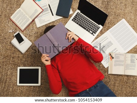 Exhausted student girl lying on the floor among textbooks, tests and gadgets, copy space. Woman covering face with book, got tired while preparing for exams. Education and overworking concept