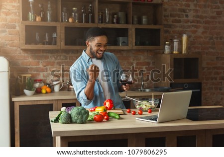 Happy african-american man watching football on laptop and drinking wine while cooking healthy food in the loft kitchen at home on sunny day. Preparing dinner for winning team celebration