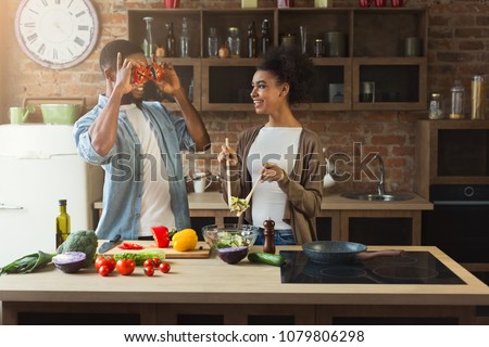 Happy african-american couple cooking healthy food and having fun together in their loft kitchen at home. Woman and man drinking wine. Preparing vegetable salad.