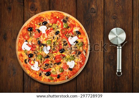 Delicious italian vegetarian pizza with tomatoes, mushrooms, peppers, corn and black olives - thin pastry crust at wooden table background with stainless steel cutter, above view