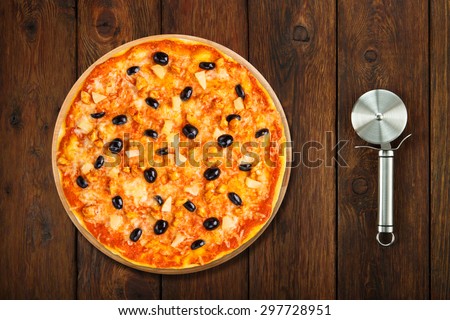 Delicious italian pizza with pineapple, chicken and black olives - thin pastry crust isolated at wooden background with stainless steel cutter, above view