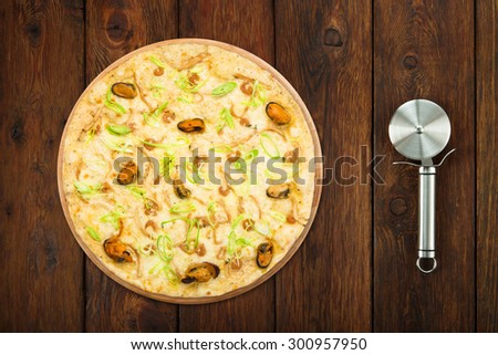 Delicious seafood pizza with shrimps, mussel, olives and leek - thin pastry crust at wooden table background with stainless steel cutter, above view