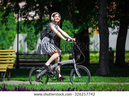 Teen student girl or young woman in plaid dress rides a folding bicycle and listens music in white earphones in the park