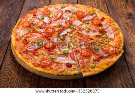 Delicious pizza with red and green hot chili peppers, bacon and cherry tomatoes - thin pastry crust at wooden background, one piece cut off