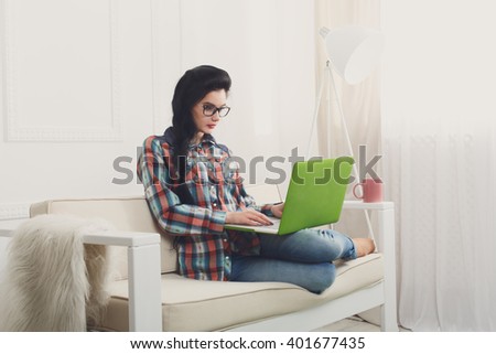 Young girl in protective computer glasses on a white sofa with laptop. Hipster freelancer woman works at home. Home office, freelance. Brunette with laptop in modern interior at white couch. Soft tone