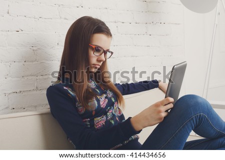 Teenager girl relax home, sitting on sofa with tablet computer. Child wears glasses, eyeglasses. Girl in eyewear with gadget tablet pc sits on sofa. Adolescence, internet addiction. High key