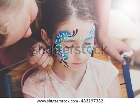 Child animator, artist's hand draws face painting to little girl. Child with funny face painting. Painter makes blue butterfly at girl's face. Children holiday, event, birthday party, entertainment.