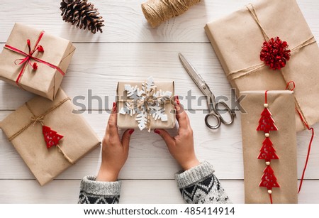 Creative hobby. Woman\'s hands show christmas holiday handmade present in craft paper with twine ribbon. Making bow at xmas gift box, decorated with snowflake. Scissors on white wooden table, top view.