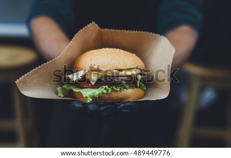 Fresh burger cooked at barbecue outdoors in craft paper. Cookout american bbq food. Big hamburger with steak meat and vegetables closeup with chef unfocused at background. Street food, fast food.