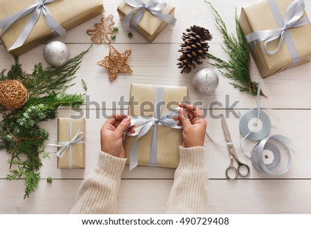 Creative diy hobby. Making bow on modern handmade christmas present, box in stylish paper with satin silver ribbon. Top view of hands on white wood table with fir tree branches, decoration of gift.