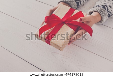 Woman\'s hands give wrapped christmas or other holiday handmade present in paper with red ribbon. Present box, decoration of gift on white wooden table with copy space