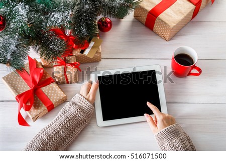 Christmas online shopping top view on wood. Female buyer touch screen of tablet, copy space. Woman has coffee, buys presents near xmas tree, among gift boxes. Winter holidays sales background