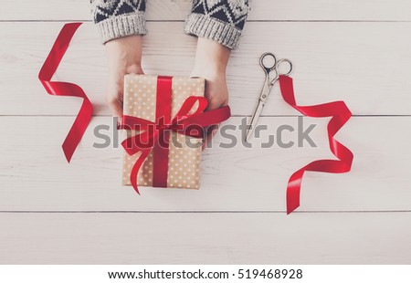Woman\'s hands give wrapped christmas or other holiday handmade present in paper with red ribbon. Present box, decoration of gift on white wooden table, top view with copy space