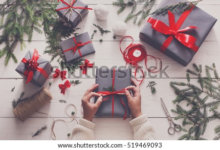 Creative hobby. Gift wrapping. Packaging modern christmas present boxes in stylish gray paper with satin red ribbon. Top view of hands on white wood table with fir tree branches, decoration