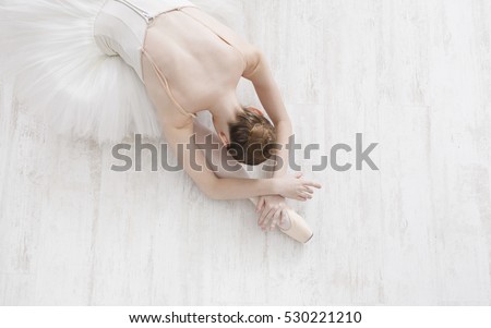 Beautiful graceful young ballerina in pointe shoes at white wooden floor background, top view from above with copy space. Ballet practice and stretching. Back of ballet dancer.