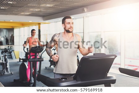 Young happy handsome man with tattoo in fitness club. Cardio workout, running on treadmill. Healthy lifestyle, guy and woman training in gym. Flare effect
