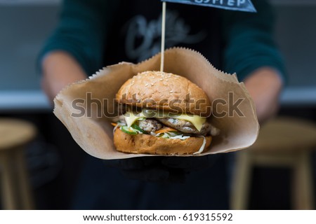 Fresh burger cooked at barbecue outdoors in craft paper. Big hamburger with steak meat and vegetables closeup, chef unfocused at background. Street fast food.