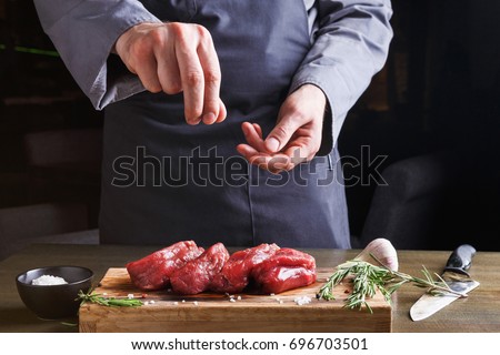 Man sprinkles filet mignon steaks with pepper salt. Chef working at open restaurant kitchen. Fresh meat, garlic and rosemary on wooden board. Modern restaurant cuisine backgroung