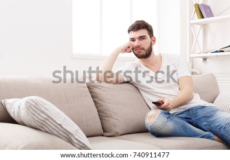 Young man watching television, using remote control to switch channels. Guy bored with what he sees on TV screen. Sitting on couch in living room at home, copy space