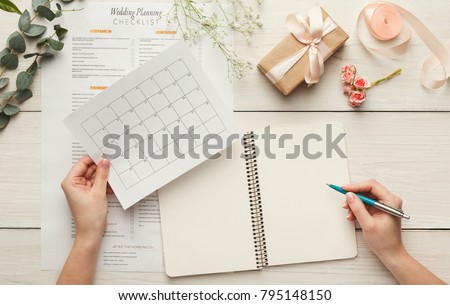 Wedding background with checklist and calendar. Female hands arranging marriage, filling in planners on white wooden table with lots of tender bridal stuff, top view