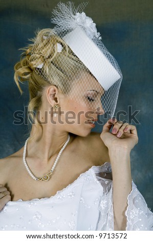 Portrait of the smiling blonde in white wedding dress and hat with veil. Studio photo
