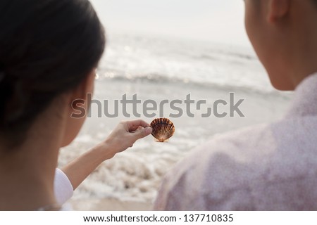 Young couple standing and looking at seashell on the beach, over the shoulder view