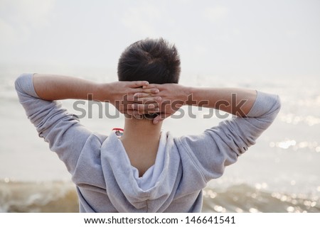 Young man with hands behind head, looking out to sea