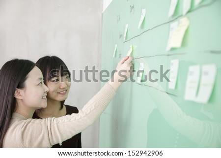 Businesswoman putting a stick note on the board, another businesswoman looking at the board