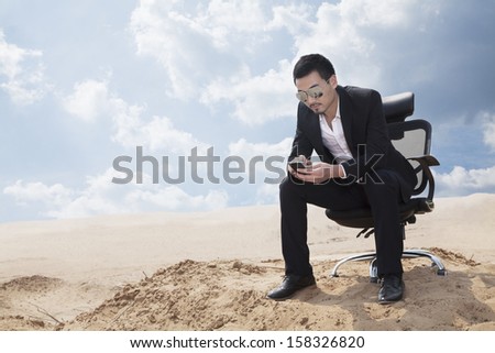 Young businessman in sunglasses sitting in office chair in desert, checking his phone