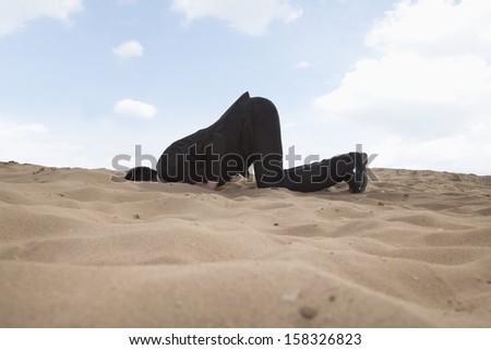 Young businessman kneeling with his head in hole in sand