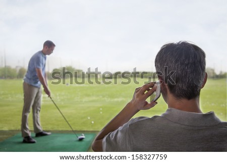 Rear view of man on phone while another man plays golf in background