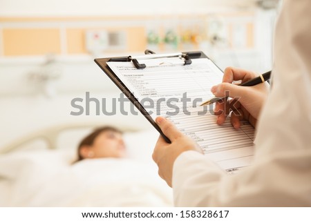 Close up of doctor writing on a medical chart with patient lying in a hospital bed