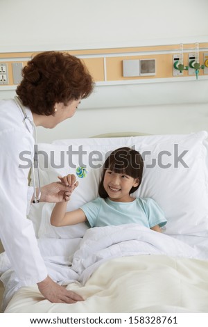 Female doctor giving lollipop to young girl lying in hospital bed
