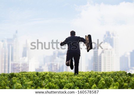 Businessman running towards the city with  briefcase in green field with plants