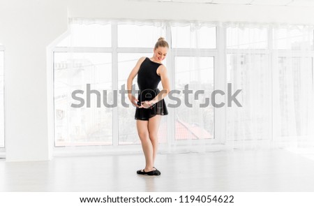 Beautiful ballerina in black leotard practising ballet moves in white bright room