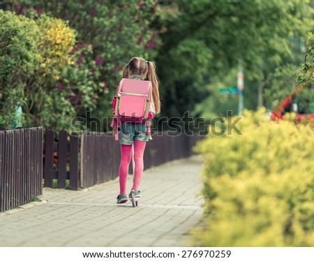 Little girl with a backpack goes to school on a scooter. back view