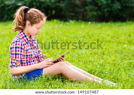 Beautiful teenager girl in casual clothes sitting on the grass with digital tablet on her knees, reading and surfing, outdoor portrait
