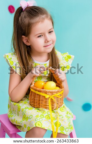 Thoughtful little girl with long blond hair wearing pink bow and holding wicker basket with yellow eggs and ribbon sitting on pink chair. Easter celebrations. Turquoise background. Studio portrait