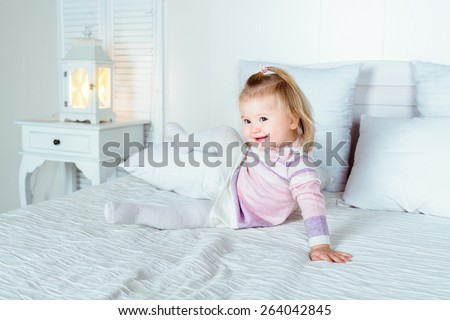 Funny and cute blond little smiling girl playing on bed in bedroom. White interior, big bed, bedside table and night lamp.