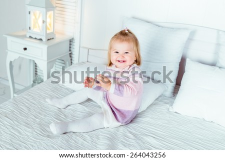 Funny and cute blond little laughing girl playing on bed in bedroom. White interior, big bed, bedside table and night lamp