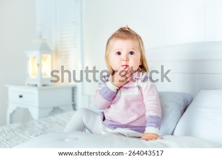 Funny amazed blond little girl sitting on bed in bedroom. White interior, big bed, bedside table and night lamp. Girl touches her mouth by hand