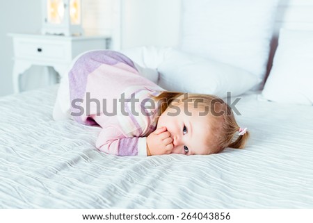 Cute little blonde girl on hands and knees on white bed in bedroom. White interior, big bed, bedside table and night lamp