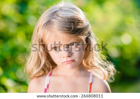 Close-up portrait of capricious blond little girl with pursed lips. Sunny summer day in beautiful park