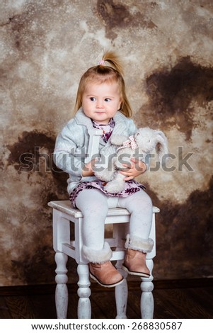 Displeased blond little girl sitting on white chair and holding her toys. Studio portrait on brown grunge background