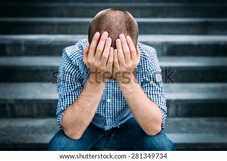 Outdoor portrait of sad young man covering his face with hands sitting on stairs. Selective focus on hands. Sadness, despair, tragedy concept
