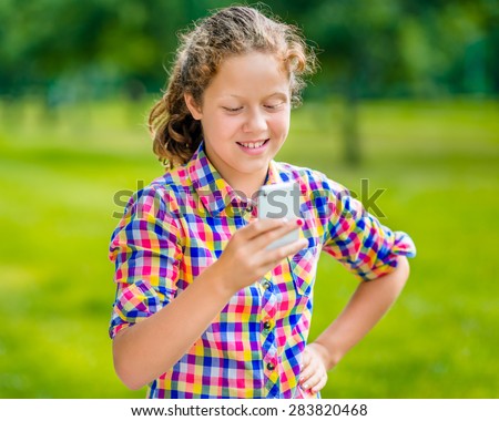 Lovely smiling teenage girl in casual clothes with smartphone in her hand, looking at screen, reading a message, using Facebook in sunny day in summer park